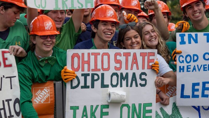 Image of Notre Dame fans decked out in green and orange, holding signs and smiling